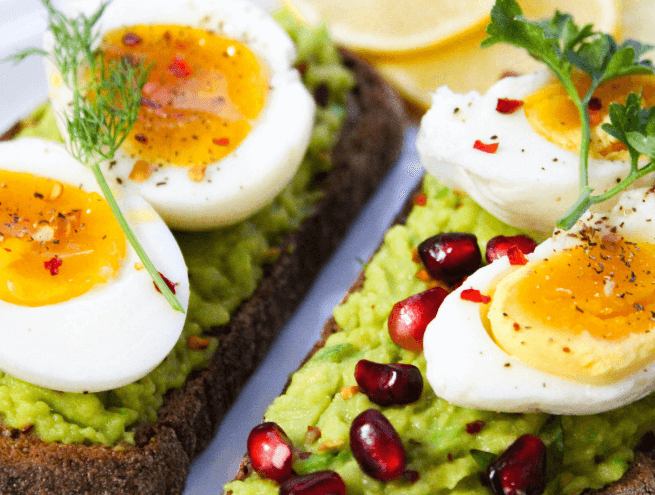 Tartines de pain complet garnies de purée d’avocat, œufs mollets coupés en deux, assaisonnés de poivre et piment, décorées avec de l’aneth, du persil et des graines de grenade.