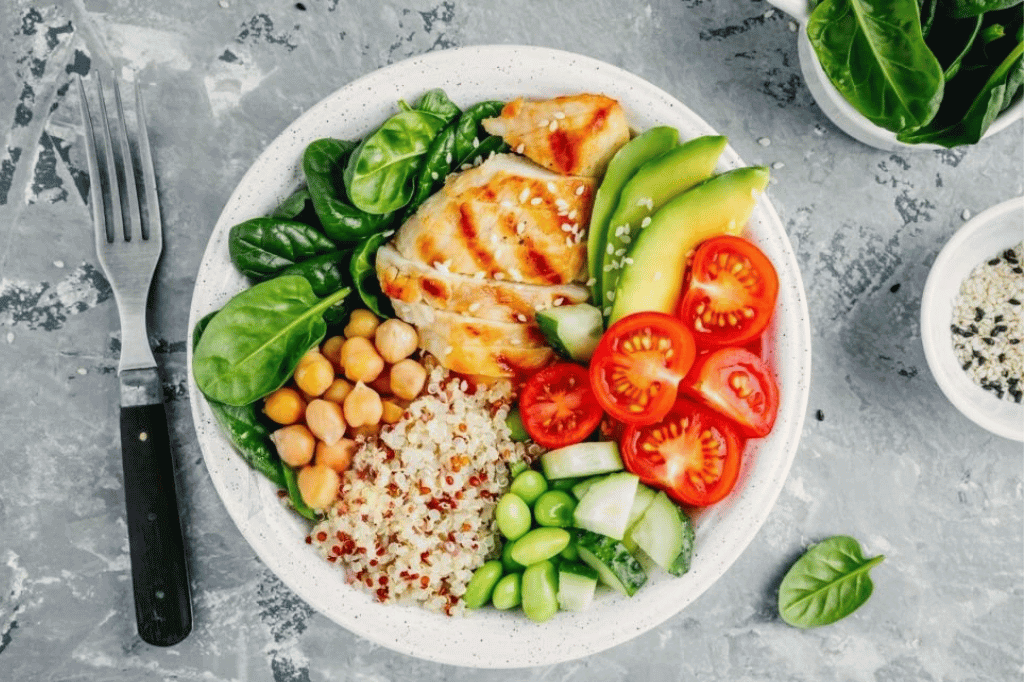 Bol équilibré composé de quinoa, pois chiches, poulet grillé, avocat, tomates cerises, concombre, épinards et edamame, représentant un repas sain et nutritif.