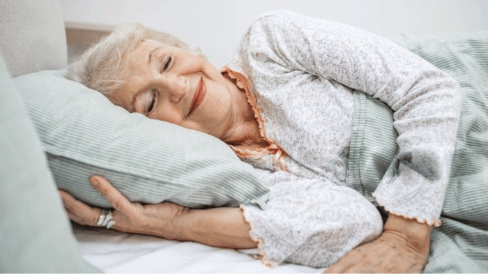 Femme âgée souriante allongée dans son lit, profitant d’un sommeil réparateur, symbole de bien-être et de repos chez les seniors.