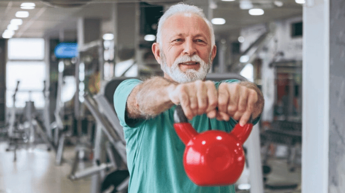 Homme senior en salle de sport tenant une kettlebell rouge, illustrant l’importance de l’exercice physique pour la santé après 60 ans.