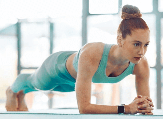 Femme en tenue de sport turquoise faisant la planche sur les avant-bras dans une salle lumineuse, concentrée sur son exercice.