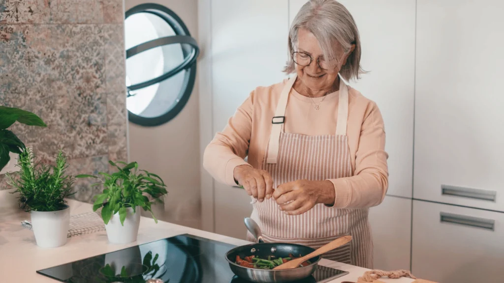 Femme senior souriante en cuisine, portant un tablier rayé et préparant des légumes dans une poêle, entourée de pots de plantes aromatiques.